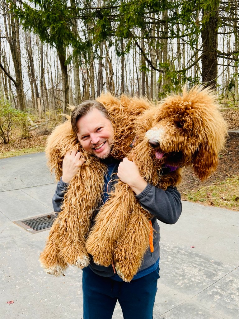 Troy Johnson, therapist at Queer Talk Therapy, with his golden doodle, Miss Betty, outdoors in a serene park, reflecting warmth and inclusivity.