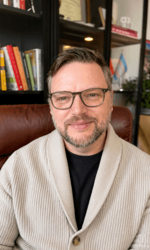 Troy Johnson, LCSW, seated in his Chicago Loop therapy office wearing a cream cardigan and glasses, smiling warmly in front of bookshelves and framed credentials at Queer Talk Therapy.