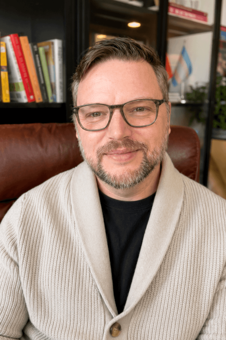 Troy Johnson, LCSW, seated in his Chicago Loop therapy office wearing a cream cardigan and glasses, smiling warmly in front of bookshelves and framed credentials at Queer Talk Therapy.